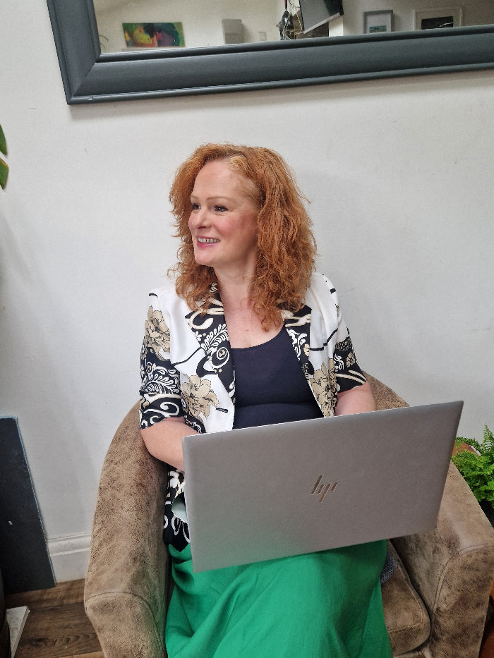 A woman with curly red hair sits in a brown armchair, smiling while using a silver HP laptop. She is wearing a black and white patterned cardigan over a dark top, with a bright green skirt or dress visible.