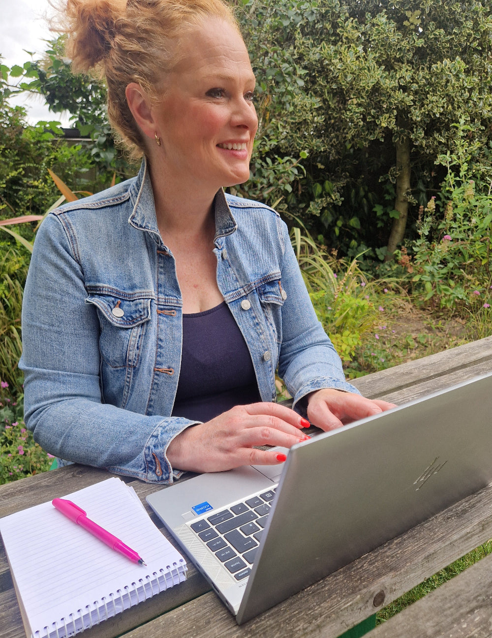 A woman with blonde hair in a bun sits at an outdoor wooden table, smiling while using a silver laptop. She is wearing a light blue denim jacket over a dark top.