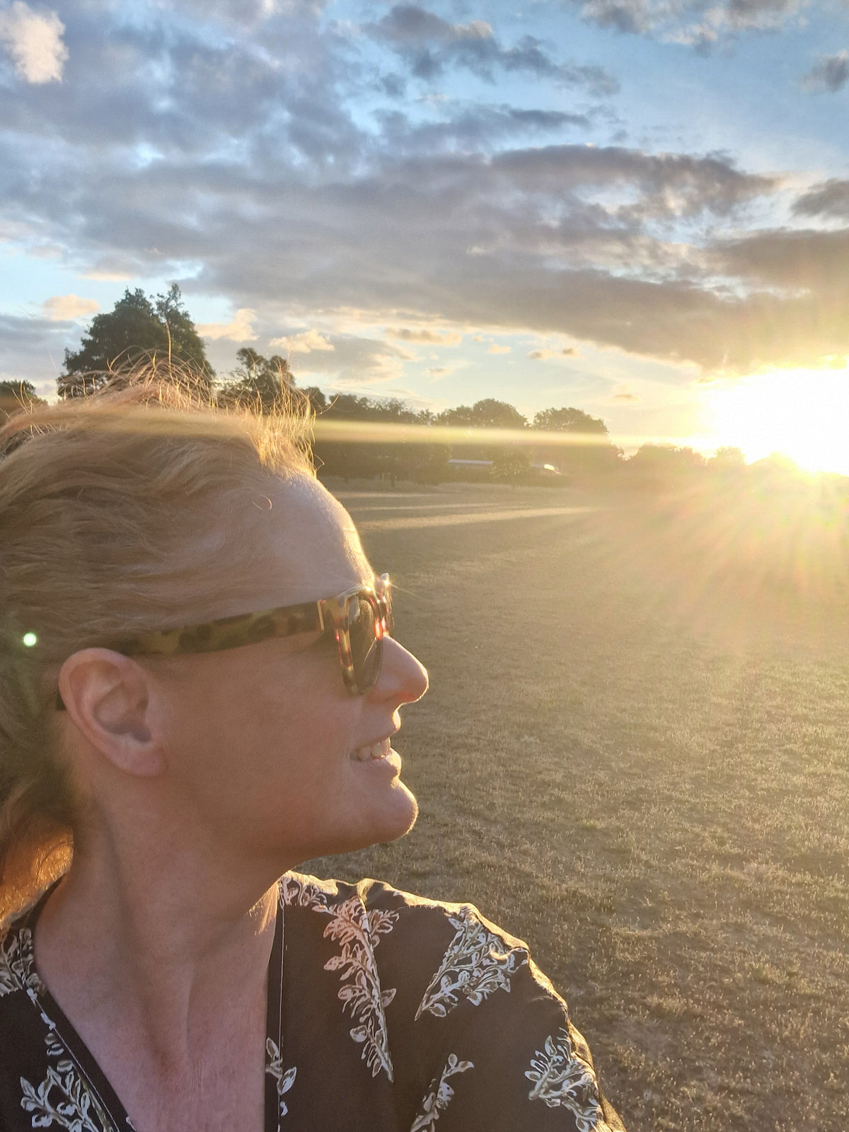 A woman outdoors at sunset wearing tortoiseshell sunglasses and a dark patterned top, with bright sun flare and clouds in the background.